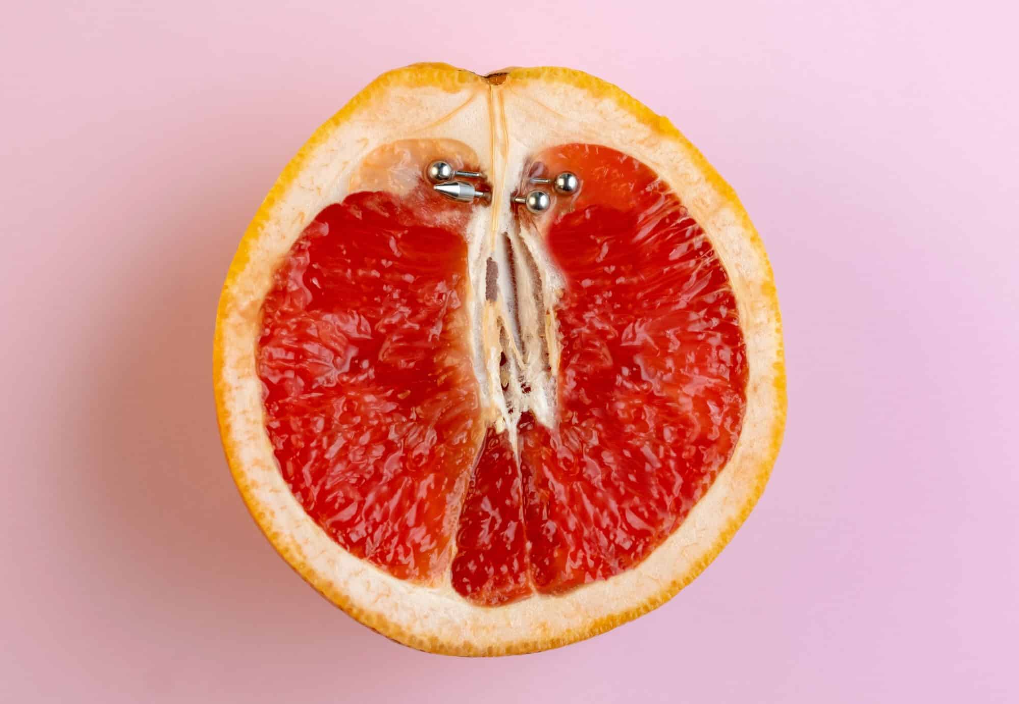 Half grapefruit with vertical barbell piercing on a pink background.