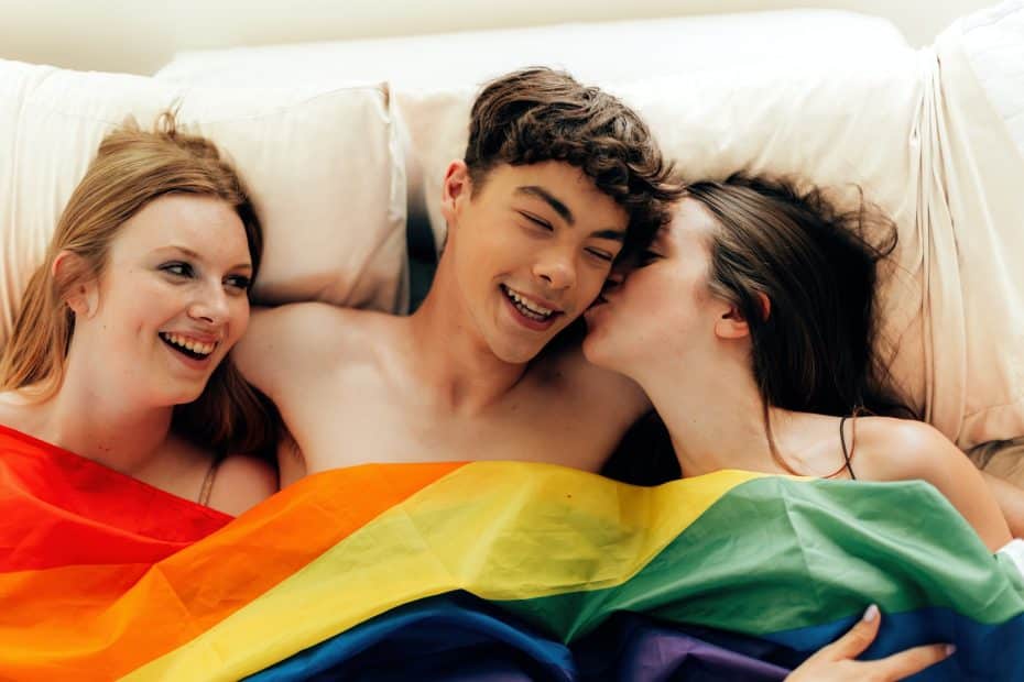 Trekantsex: To jenter og en ung mann ligger i en seng med et LHBT flagg over sengen. Illustrerer at de skal ha trekantsex.