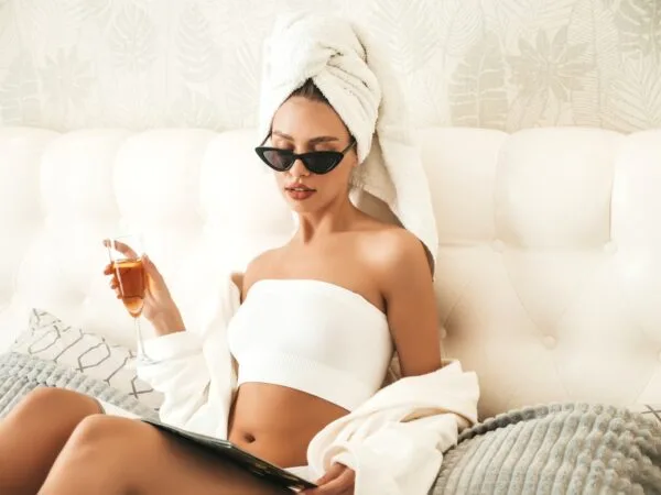 Ung flott dame sitter i sengen på et hotellrom i syden. I hånden holder hun et glass med vin.