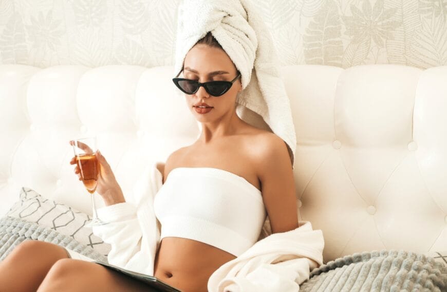 Ung flott dame sitter i sengen på et hotellrom i syden. I hånden holder hun et glass med vin.