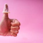 Rosa bakgrunn, mannlig hånd som gir "thumbs up" med en kondom tredd ned over tommelen. Illustrasjon til artikkel om seksuelt samtykke.