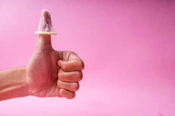 Rosa bakgrunn, mannlig hånd som gir "thumbs up" med en kondom tredd ned over tommelen. Illustrasjon til artikkel om seksuelt samtykke.